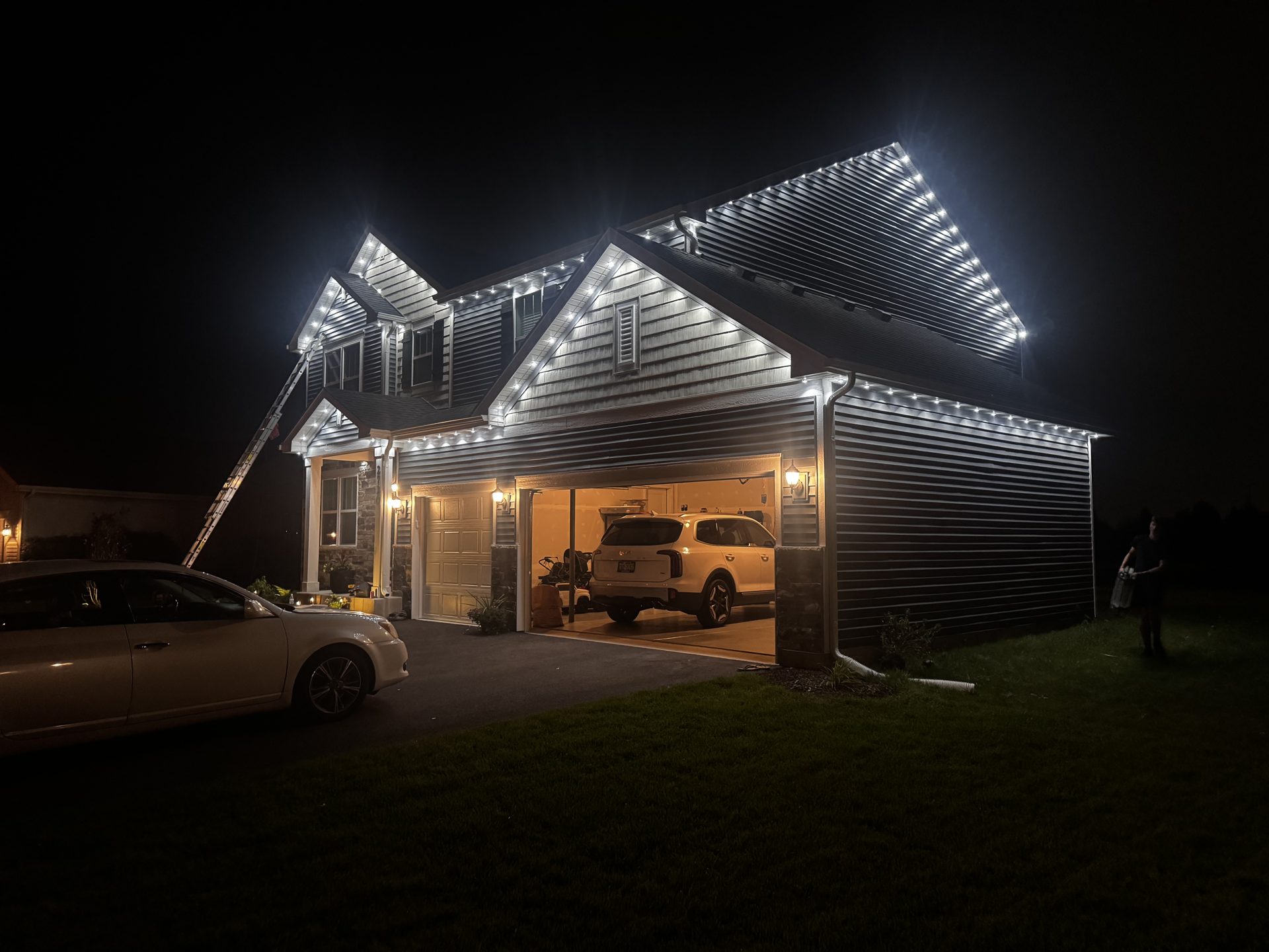 Two-story home at night with permanent cool white lighting along the roofline, gables, and garage